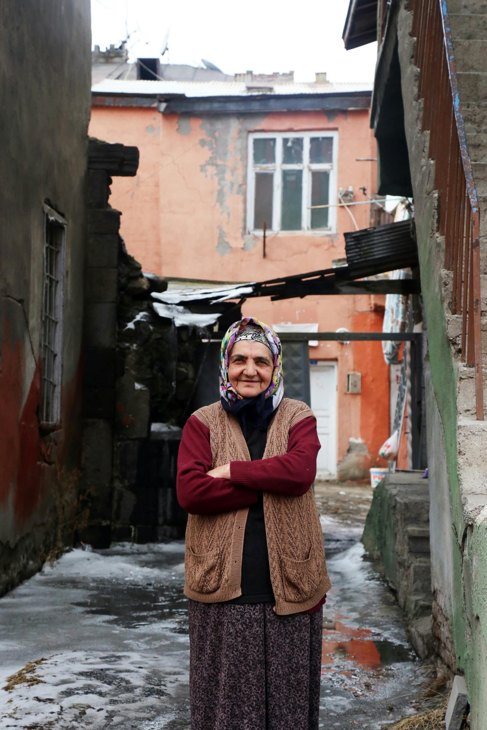 Portrait of an elderly woman in a vibrant headscarf, smiling in a rustic urban alley.